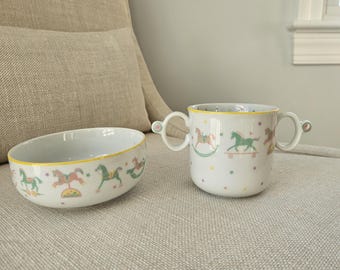 Puede incluir: Un cuenco y una taza de dos asas de cerámica blanca con un patrón repetitivo de caballos balancín de colores y pequeñas estrellas. Ambas piezas tienen un borde amarillo fino y la taza incluye pequeños detalles en las asas.