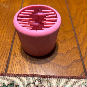 May include: A pink, cylindrical object with a series of horizontal, parallel slots. The object is made of a soft material and has a rounded top. The item is sitting on a wooden surface.