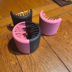May include: Three hair styling diffusers in the image. One is black, and the other is pink. The third diffuser is split, with one half pink and the other black. Each diffuser has a cylindrical base with a comb-like top.