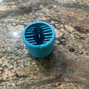 May include: A light blue plastic hair dryer diffuser with a round shape and a grid of holes. The diffuser is split in half and is sitting on a countertop.