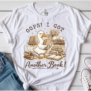 Könnte beinhalten: Weißes T-Shirt mit einer Illustration einer Ente, die ein Buch hält, umgeben von Bücherstapeln und dem Text "OOPS! I GOT Another Book!". Das Design hat eine Vintage-Ästhetik mit braunen und beigen Farbtönen.
