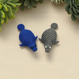 Puede incluir: Dos figuras con forma de foca. Una es azul, la otra gris. Ambas tienen un patr&oacute;n texturizado, similar a escamas, en la espalda y peque&ntilde;as aletas. Las figuras est&aacute;n sobre un fondo claro con vetas de madera, con suculentas verdes en las esquinas.