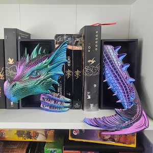 May include: A pair of dragon-themed bookends in iridescent colors of teal, purple, and green. One bookend is the dragon's head, the other is the tail. The bookends are displayed on a white shelf with books.