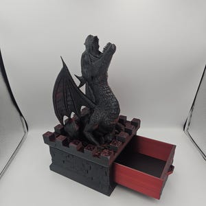 May include: A black dragon figurine with open mouth and detailed scales sits atop a black and red box with an open drawer. The dragon has large wings and is posed as if roaring. The box has a castle-like design.