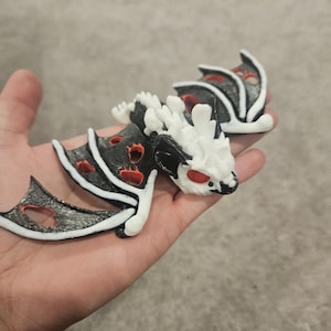 May include: A pair of dragon figurines, each featuring a black body with red accents and white detailing. The dragons have red eyes and are held in a hand. The wings are black with a silver outline.