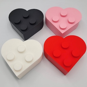 May include: Four heart-shaped boxes in black, pink, white, and red. Each box has four raised circular studs on the top, resembling building blocks. The boxes are arranged on a white surface.