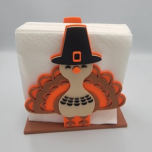 May include: A Thanksgiving-themed napkin holder featuring a turkey design. The turkey has a black pilgrim hat, orange and brown feathers, and a beige body. The holder is made of brown material and holds a stack of white napkins.