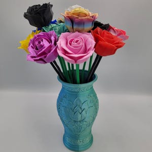May include: A teal vase with a floral design holds a bouquet of colorful roses. The roses are in shades of pink, red, yellow, and black. The stems are green and black, creating a vibrant and eye-catching arrangement.