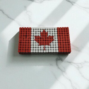 Pode incluir: Um objeto retangular que se assemelha &agrave; bandeira canadense, composto por pequenos quadrados vermelhos, brancos e pretos. A bandeira apresenta uma folha de bordo vermelha no centro. O objeto est&aacute; sobre uma superf&iacute;cie de m&aacute;rmore branco.