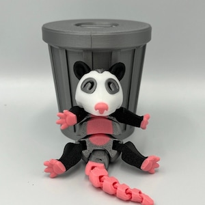 Op de afbeelding: Een 3D-geprint opossum-speelgoed met een roze staart, voeten en neus, zwarte armen en oren en een wit gezicht. Het speelgoed staat voor een grijze vuilnisbak. Het lichaam van de opossum is grijs en roze.