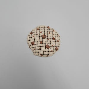 May include: A round, off-white ceramic coaster with a grid pattern and brown accents. The coaster has a cookie-like appearance, with brown spots resembling chocolate chips. The design is simple and minimalist.