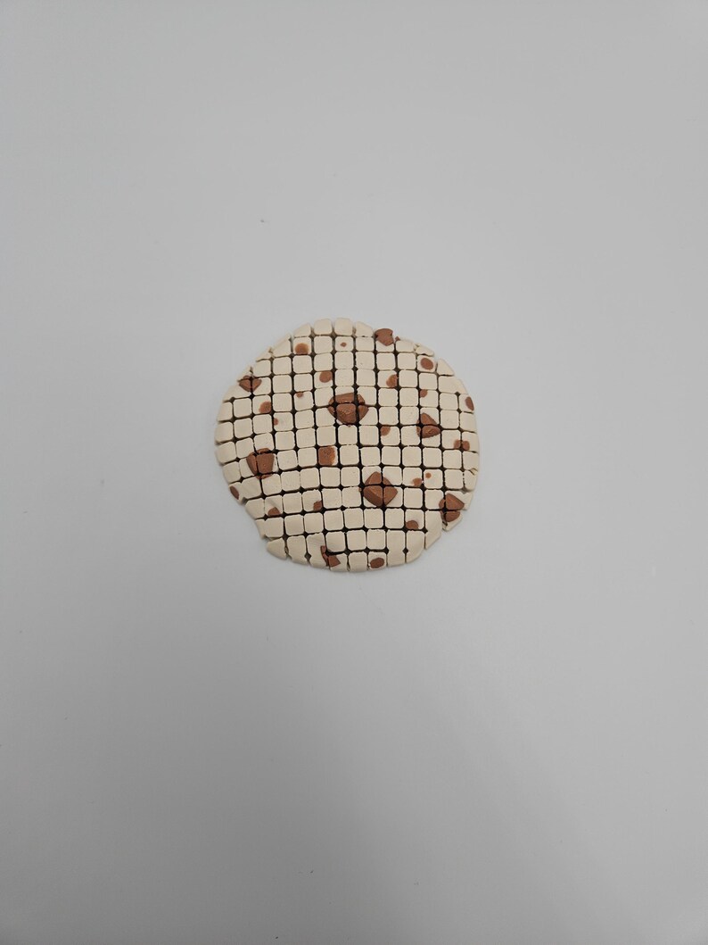 May include: A round, off-white ceramic coaster with a grid pattern and brown accents. The coaster has a cookie-like appearance, with brown spots resembling chocolate chips. The design is simple and minimalist.