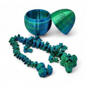 May include: A teal and green mushroom-shaped container with a lid, open to reveal a string of teal and green mushroom-shaped beads. The beads are connected by a thin string and have a textured surface.