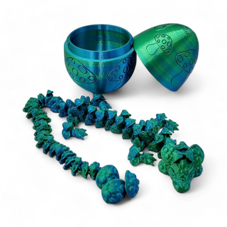 May include: A teal and green mushroom-shaped container with a lid, open to reveal a string of teal and green mushroom-shaped beads. The beads are connected by a thin string and have a textured surface.