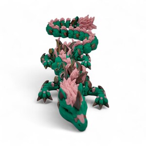 May include: A green and pink articulated dragon figurine. The dragon has a detailed design with a green body, pink accents, and brown details. The dragon is posed on a white surface.