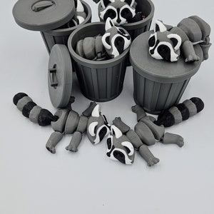 May include: Gray 3D-printed raccoon toys with black and white markings are scattered around small gray trash cans. Some raccoons are inside the cans, while others are disassembled nearby. The trash cans have lids, and the background is white.