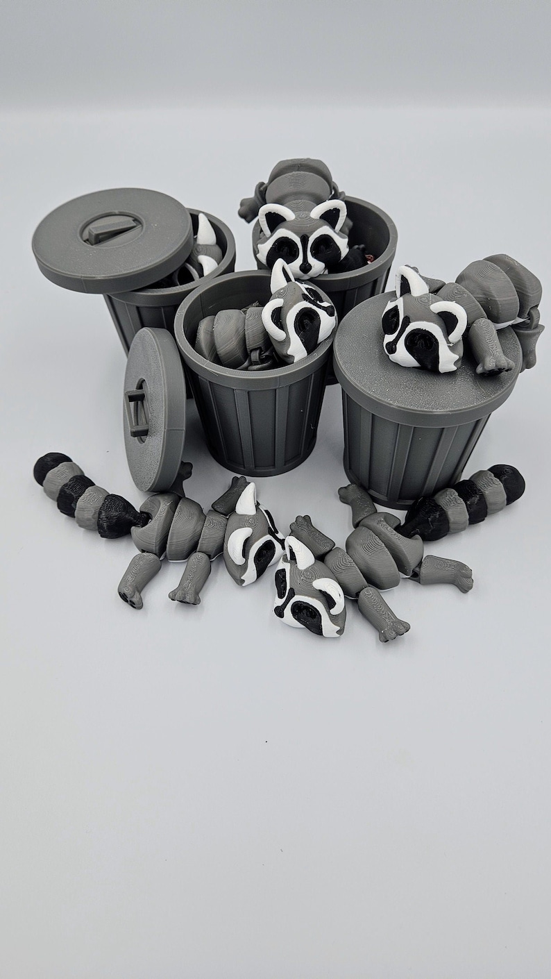 May include: Gray 3D-printed raccoon toys with black and white markings are scattered around small gray trash cans. Some raccoons are inside the cans, while others are disassembled nearby. The trash cans have lids, and the background is white.