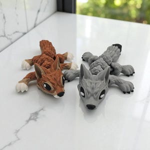 Peut inclure: Deux figurines d'animaux : un &eacute;cureuil brun et un loup gris. L'&eacute;cureuil a des pattes blanches et un ventre blanc. Le loup a une pointe noire sur sa queue. Les deux sont allong&eacute;s, les pattes tendues.