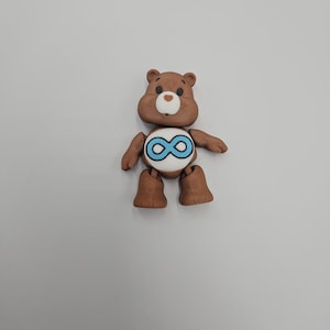 May include: A brown bear figurine with a white belly and a blue infinity symbol. The bear has a light brown snout and a small, round, white nose. The bear's arms and legs are also brown.