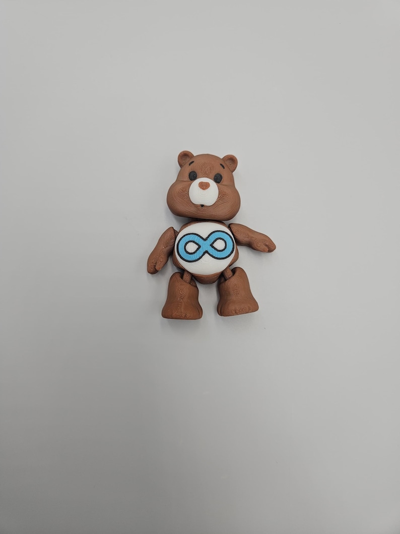 May include: A brown bear figurine with a white belly and a blue infinity symbol. The bear has a light brown snout and a small, round, white nose. The bear's arms and legs are also brown.