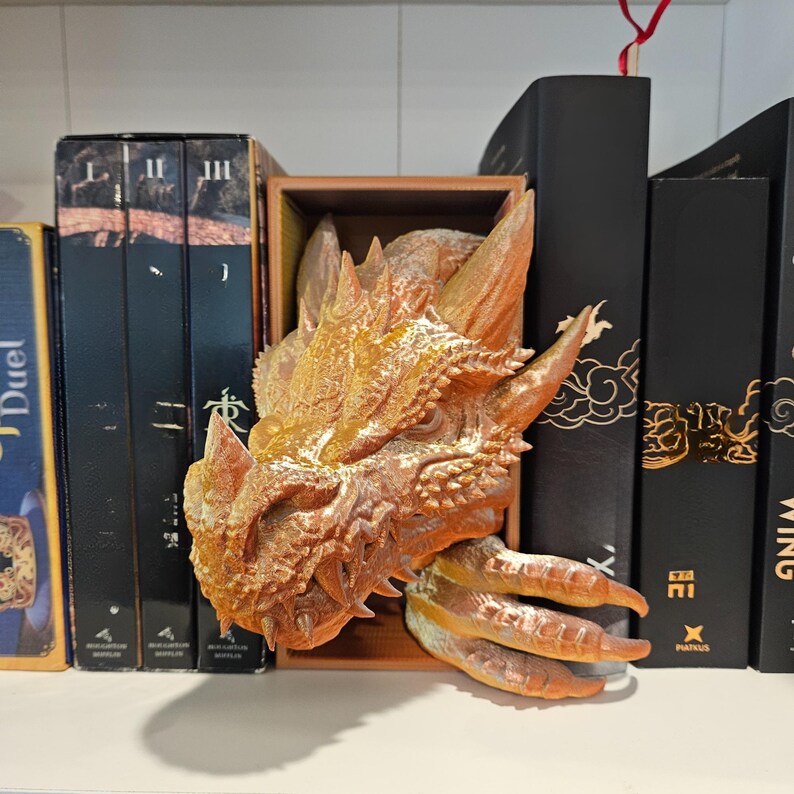 May include: A detailed dragon head bookend in shades of gold and brown, with textured scales and sharp claws. The dragon head is positioned within a wooden box, designed to hold books. The bookend is placed among a collection of black books.