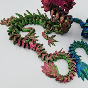 May include: A detailed, articulated dragon sculpture in shades of green, pink, and blue. The dragon features a textured, scale-like design and is posed on a white surface. The dragon's head and body are intricately designed.