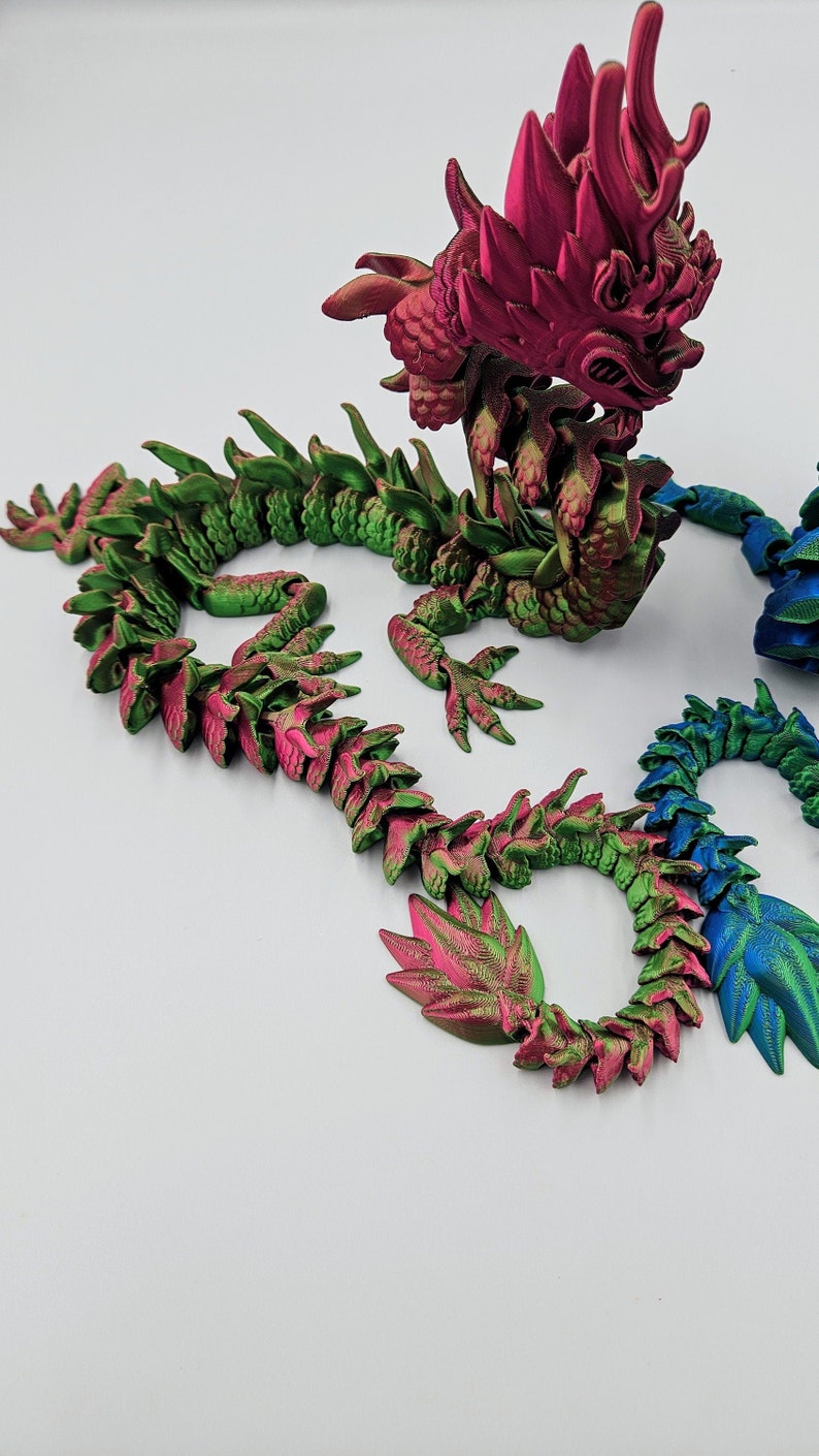 May include: A detailed, articulated dragon sculpture in shades of green, pink, and blue. The dragon features a textured, scale-like design and is posed on a white surface. The dragon's head and body are intricately designed.