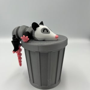 Op de afbeelding: Een grijze 3D-geprinte opossum-figuur rustend op een bijpassende grijze vuilnisbak. De opossum heeft een wit gezicht, een roze neus en poten en een gestreept lichaam. De vuilnisbak heeft verticale lijnen.