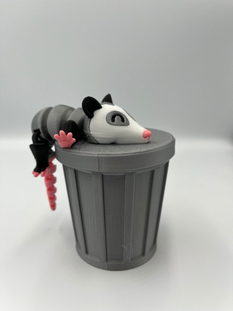 Op de afbeelding: Een grijze 3D-geprinte opossum-figuur rustend op een bijpassende grijze vuilnisbak. De opossum heeft een wit gezicht, een roze neus en poten en een gestreept lichaam. De vuilnisbak heeft verticale lijnen.