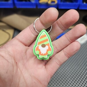 Pode incluir: Um chaveiro de gnomo verde e laranja com barba branca e chap&eacute;u listrado. O chaveiro est&aacute; preso a um anel prateado.