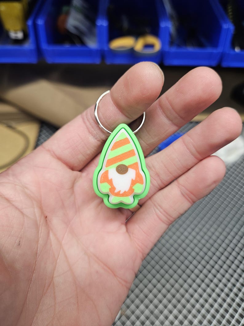 Pode incluir: Um chaveiro de gnomo verde e laranja com barba branca e chap&eacute;u listrado. O chaveiro est&aacute; preso a um anel prateado.