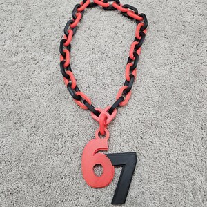 May include: A necklace with a chain of alternating black and red links. A red pendant with the number 6 and a black pendant with the number 7 are attached to the chain. The necklace is lying on a gray surface.