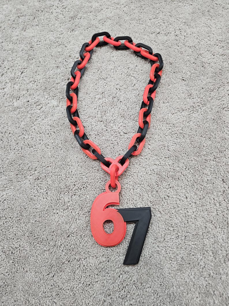 May include: A necklace with a chain of alternating black and red links. A red pendant with the number 6 and a black pendant with the number 7 are attached to the chain. The necklace is lying on a gray surface.