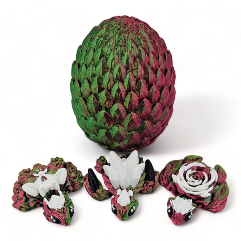 Puede incluir: Un huevo de drag&oacute;n verde y rojo con tres peque&ntilde;as figuras de drag&oacute;n. Las figuras de drag&oacute;n son verdes, rojas y blancas con un dise&ntilde;o de rosa.
