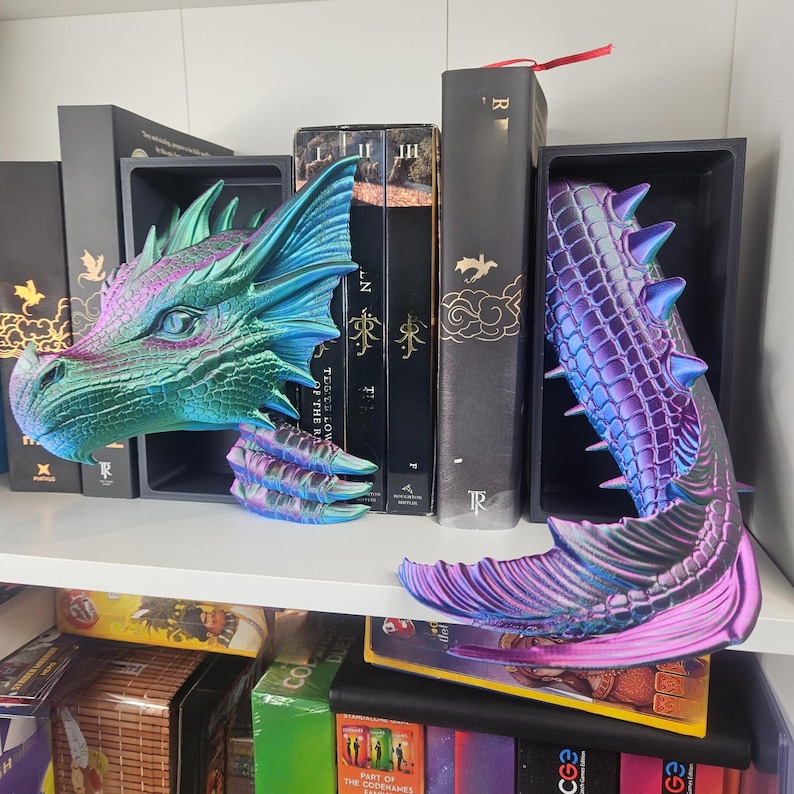 May include: Dragon-themed bookends in iridescent teal, purple, and blue. One bookend is a dragon head, the other a tail. The bookends are on a white shelf with books in the background. The bookends are made of a 3D printed material.