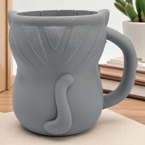 May include: A gray ceramic mug shaped like a cat, featuring a handle, ears, and a tail. The mug is designed with a textured surface and is suitable for beverages. The mug is sitting on a beige surface.