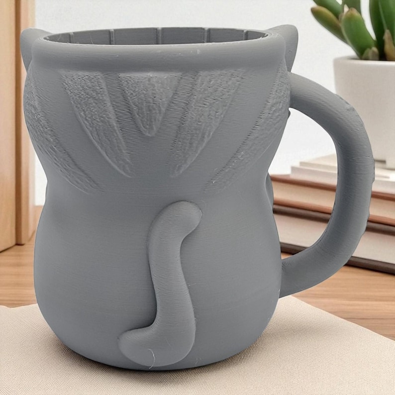 May include: A gray ceramic mug shaped like a cat, featuring a handle, ears, and a tail. The mug is designed with a textured surface and is suitable for beverages. The mug is sitting on a beige surface.