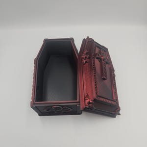 May include: An open, red decorative coffin with ornate detailing. The interior is black. The lid is open, revealing the inside. The coffin is made of a dark red material with decorative carvings.
