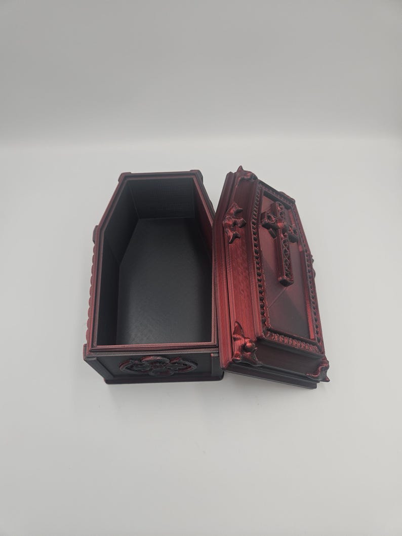 May include: An open, red decorative coffin with ornate detailing. The interior is black. The lid is open, revealing the inside. The coffin is made of a dark red material with decorative carvings.