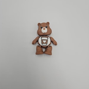 May include: A brown bear figurine with a white belly featuring a coffee cup design. The bear has a smiling face and is standing upright. The figurine is made of a smooth material and is set against a plain white background.