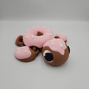 Pode incluir: Uma escultura extravagante em forma de donut, com cobertura rosa e granulado branco. A escultura tem uma base marrom e &eacute; acompanhada por pe&ccedil;as menores semelhantes, criando um design l&uacute;dico com tema de comida. O donut tem um buraco no centro.