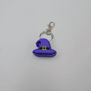 May include: A purple witch hat keychain with a black buckle and yellow trim. The keychain is attached to a silver split ring.