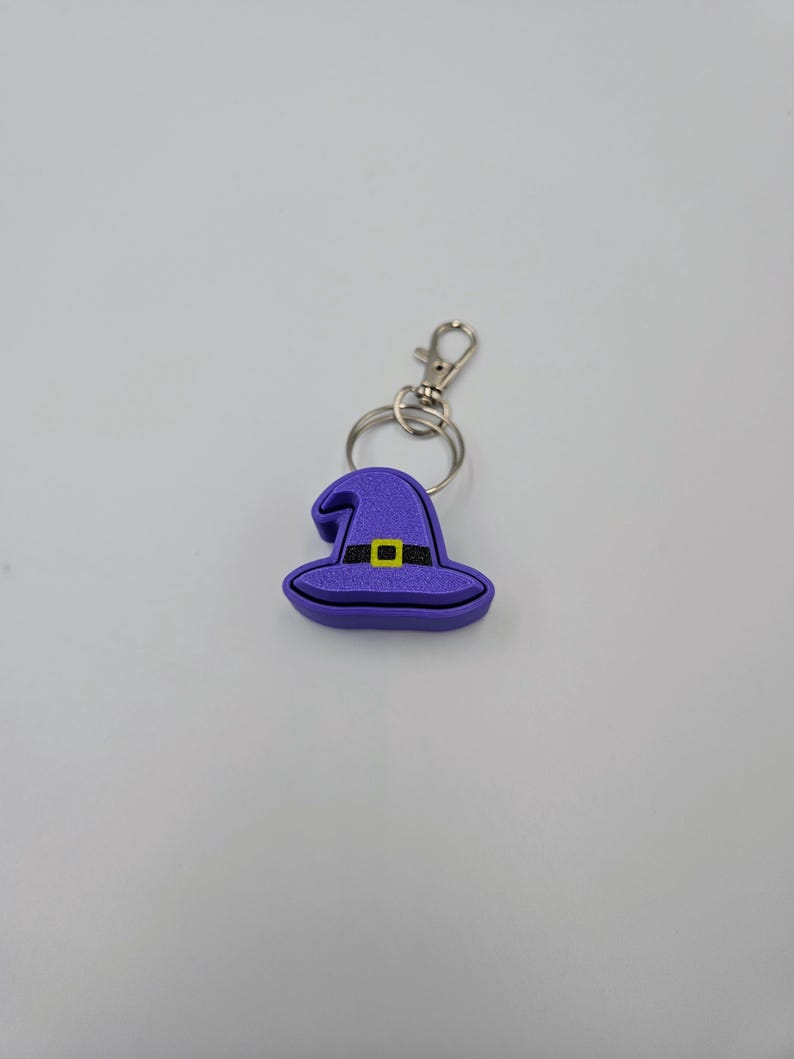 May include: A purple witch hat keychain with a black buckle and yellow trim. The keychain is attached to a silver split ring.