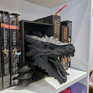 Pode incluir: Um divisor de estante em forma de drag&atilde;o preto. A cabe&ccedil;a e as garras do drag&atilde;o se estendem de uma prateleira retangular preta, com a cabe&ccedil;a posicionada para parecer que est&aacute; emergindo da prateleira. Livros s&atilde;o vis&iacute;veis em ambos os lados do drag&atilde;o.