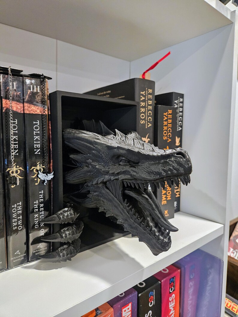 Pode incluir: Um divisor de estante em forma de drag&atilde;o preto. A cabe&ccedil;a e as garras do drag&atilde;o se estendem de uma prateleira retangular preta, com a cabe&ccedil;a posicionada para parecer que est&aacute; emergindo da prateleira. Livros s&atilde;o vis&iacute;veis em ambos os lados do drag&atilde;o.
