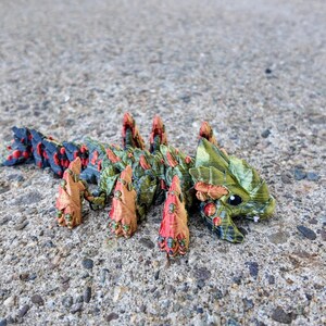 May include: A small, green, red, and black 3D printed creature with a spiky back and a large head. The creature has a large, open mouth and small, black eyes.