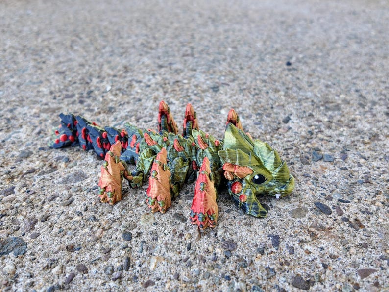 May include: A small, green, red, and black 3D printed creature with a spiky back and a large head. The creature has a large, open mouth and small, black eyes.