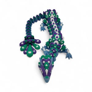 May include: A handcrafted, whimsical dragon sculpture in shades of blue, green, purple, and white. The dragon's body is intricately detailed with layered, petal-like shapes, and it has a long, curved tail. The sculpture is set against a white background.