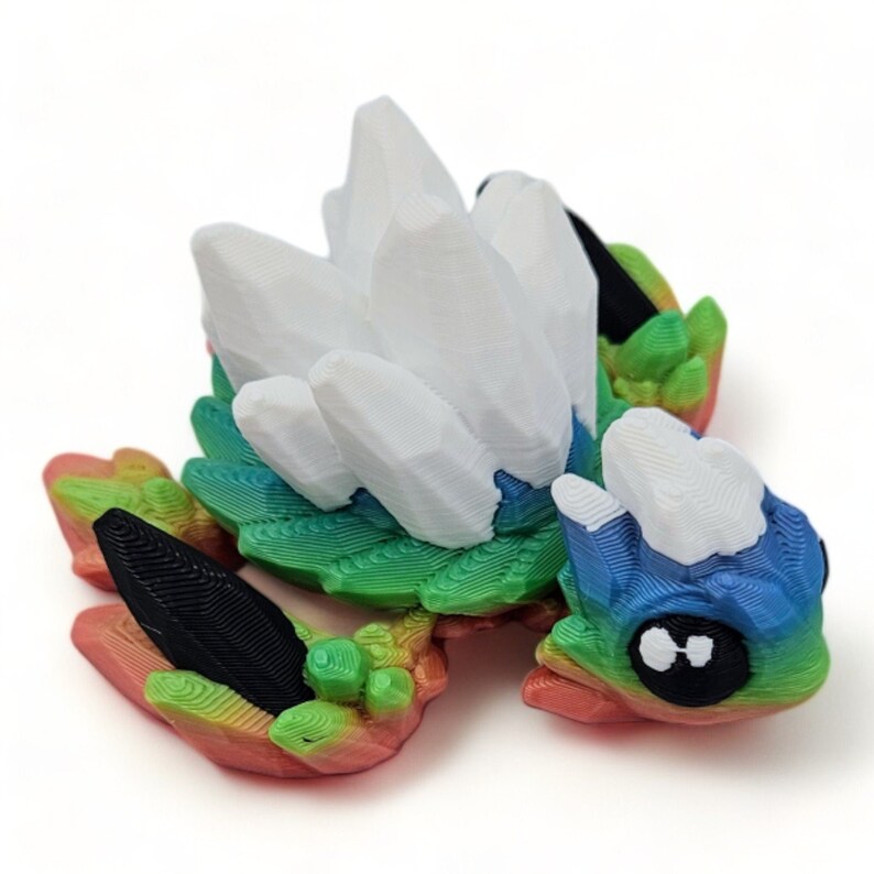 Puede incluir: Una figurita impresa en 3D de una peque&ntilde;a criatura colorida con alas de cristal blanco. La criatura tiene un cuerpo verde, azul y rojo con ojos negros.