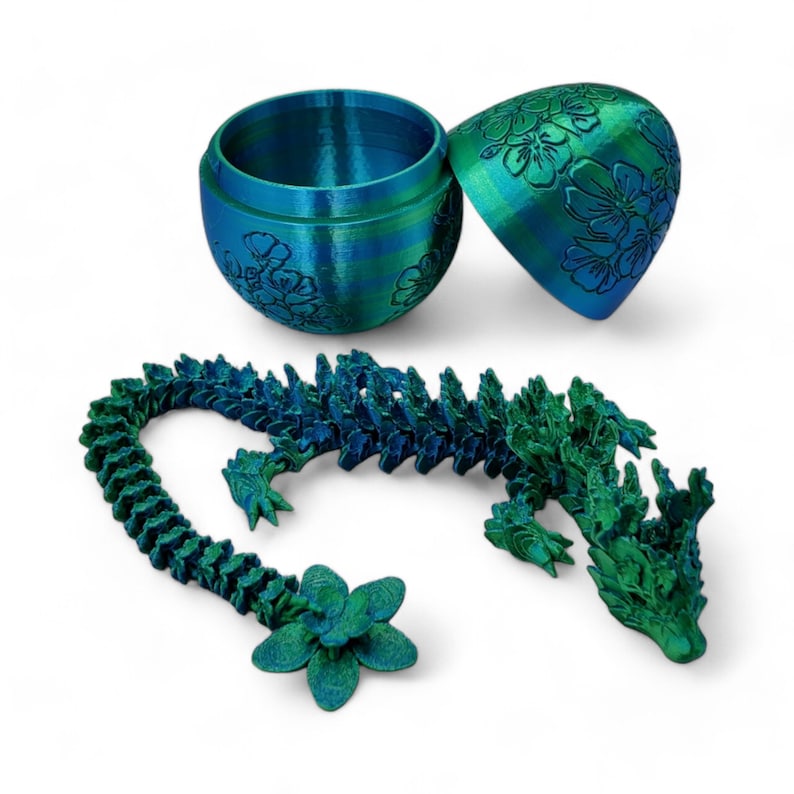 Pu&ograve; includere: Una statuetta di drago stampata in 3D di colore verde acqua e verde con un contenitore a forma di uovo con motivo floreale verde acqua e verde.